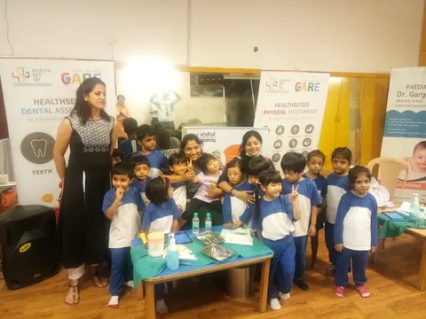 Dr Gargi Mathur standing with a group of schoolchildren in uniform at a school health assessment camp, beside health-screening banners and a table with checkup supplies.